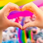 Supporting hands make heart sign and wave in front of a rainbow flag flying on the sidelines of a summer gay pride parade